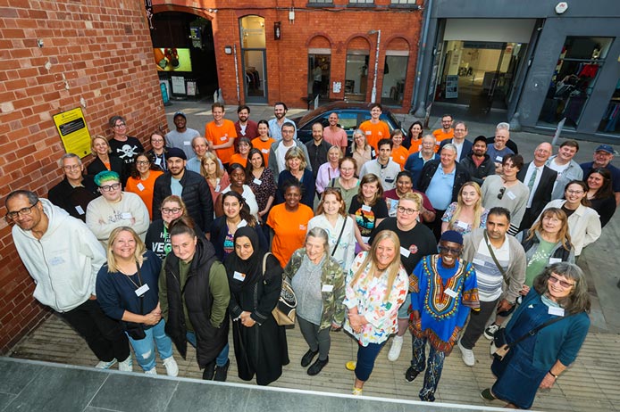 A high-angle group portrait of a diverse crowd of around fifty people smiling and posing together in an urban outdoor alleyway.