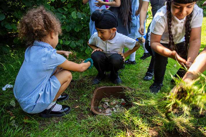 Several young school children crouch on the grass to fill a small container with plant clippings.