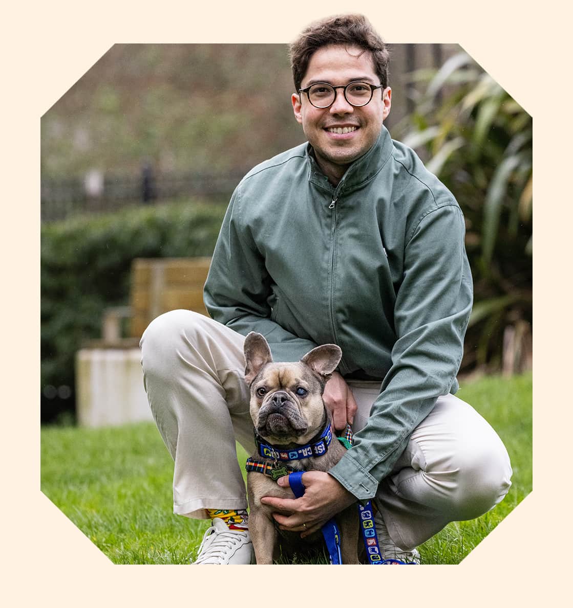 A smiling man crouches on green grass, gently holding a French bulldog with one cloudy eye.